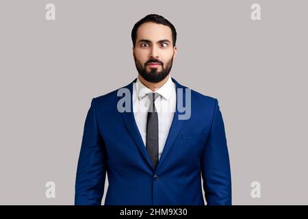 Portrait d'un jeune homme barbu adulte qui regarde les yeux croisés, qui s'amuse avec l'expression du visage stupide, qui joue à l'imbécile et porte un costume de style officiel. Prise de vue en studio isolée sur fond gris. Banque D'Images