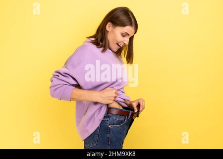 Vue latérale de la femme heureuse aux cheveux foncés montrant la perte de poids réussie, exprimant le bonheur, concept de régime, portant le pull à capuche violet. Studio d'intérieur isolé sur fond jaune. Banque D'Images