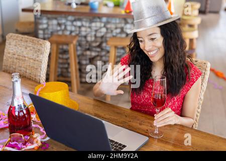 Jeune femme biraciale souriante se faisant passer la main tout en profitant de la fête sur appel vidéo sur ordinateur portable au café Banque D'Images