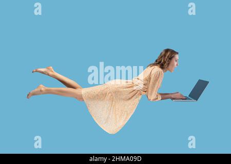 Vol stationnaire dans l'air. Surprise fille excitée robe jaune lévitant avec ordinateur portable, clavier dactylographiant, lisant des nouvelles choquantes sur l'écran d'ordinateur portable tout en volant dans les airs. studio tourné isolé sur fond bleu Banque D'Images