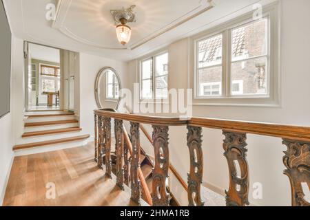 Couloir avec fenêtres et escalier en bois avec rambarde ornementale placée contre la porte ouverte de la pièce avec table à la maison avec miroir Banque D'Images