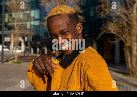 Vue latérale d'un homme afro-américain enchanté avec des cheveux teints debout sur une rue ensoleillée avec des arbres et un bâtiment moderne dans la ville pointant et regardant la came Banque D'Images