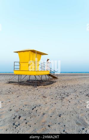 Loin non reconnaissable femme voyageur en robe et chapeau debout seul près de la cabine de sauveteur jaune situé sur la plage de sable et contemplant la mer pendant Banque D'Images