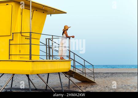 Loin non reconnaissable femme voyageur en robe et chapeau debout seul près de la cabine de sauveteur jaune situé sur la plage de sable et contemplant la mer pendant Banque D'Images