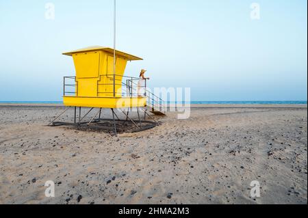 Loin non reconnaissable femme voyageur en robe et chapeau debout seul près de la cabine de sauveteur jaune situé sur la plage de sable et contemplant la mer pendant Banque D'Images