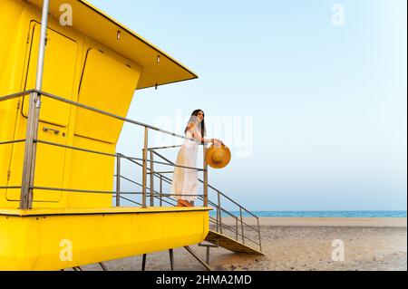 Femme voyageur en robe et chapeau debout seul près de la cabine de sauveteur jaune situé sur la plage de sable et contemplant la mer pendant les vacances d'été Banque D'Images