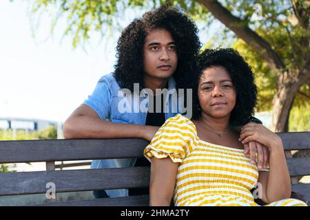 Petit ami hispanique assis avec une petite amie sur un banc en bois dans le parc tout en regardant la caméra Banque D'Images