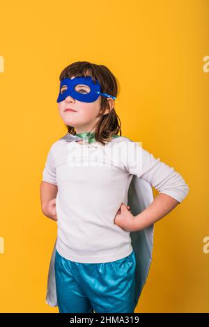 Petite fille en vêtements super-héros et masque regardant loin tout en se tenant contre fond jaune avec les mains sur la taille Banque D'Images