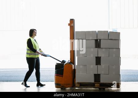 Vue latérale du corps entier de l'employé de l'entrepôt en uniforme haute vis gilet poussant spécial gerbeur manuel avec pile de boîtes en carton lourd Banque D'Images