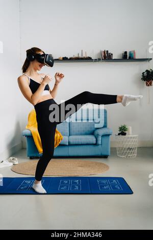 Vue latérale sur toute la longueur de la coupe anonyme femme dans les vêtements de sport effectuant des coups de pied sur le tapis lors de l'entraînement dans le casque VR à la maison Banque D'Images