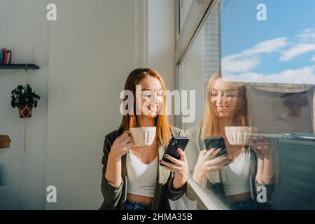 Bonne femme rouge aux cheveux debout contre la fenêtre tout en utilisant le téléphone portable et en buvant du café le jour ensoleillé Banque D'Images