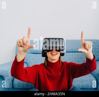 Femme dans des lunettes VR souriant et pointant vers le haut tout en étant assis contre le canapé et en explorant la réalité virtuelle Banque D'Images