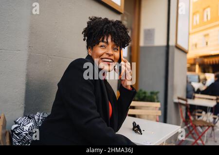 Une femme afro-américaine aux cheveux bouclés et à la mode, assise à table dans un café en plein air, qui a eu une conversation téléphonique à l'écart Banque D'Images