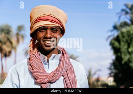 Homme arabe gai vêtu d'un turban traditionnel regardant l'appareil photo et souriant sur un fond flou de ville exotique en été Banque D'Images