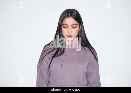 Jeune femme attrayante avec de longs cheveux noirs dans un sweat à capuche décontracté regardant l'appareil photo avec le sourire sur fond clair en studio Banque D'Images