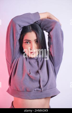 Jeune femme attrayante avec de longs cheveux noirs dans un sweat à capuche décontracté regardant l'appareil photo avec le sourire sur fond clair en studio Banque D'Images
