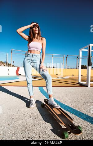 Pleine longueur de jeune brunette mince et confiante en Jean et debout avec planche à roulettes dans le parc de skate en plein soleil d'été et en regardant loin Banque D'Images