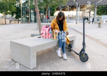 Tout le corps de la femelle focalisée dessin sur tablette avec stylet tout en étant assis sur un banc avec sac à dos près du scooter électrique en ville Banque D'Images