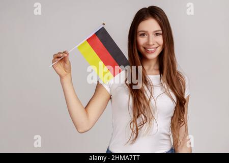 Femme attirante à cheveux foncés portant le drapeau de l'Allemagne, célébrant le jour de l'Allemagne - 3th octobre, exprimant positif, portant un T-shirt blanc. Prise de vue en studio isolée sur fond gris. Banque D'Images