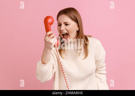 Portrait d'une femme blonde irritée en colère criant dans un combiné rouge, ayant une conversation désagréable, exprimant l'agression, portant un chandail blanc. Studio d'intérieur isolé sur fond rose. Banque D'Images