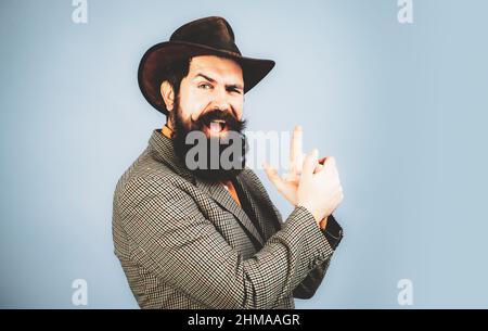 Jeune beau barbu insouciant drôle homme en costume avec les doigts comme des fusils. Gros plan de brunet fin dans un chapeau avec imagine gun dans les mains sur isolé Banque D'Images