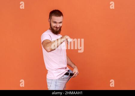 Portrait de vue latérale d'un homme barbu mince et attrayant montrant une perte de poids réussie, conception de régime, portant un T-shirt rose. Studio d'intérieur isolé sur fond orange. Banque D'Images