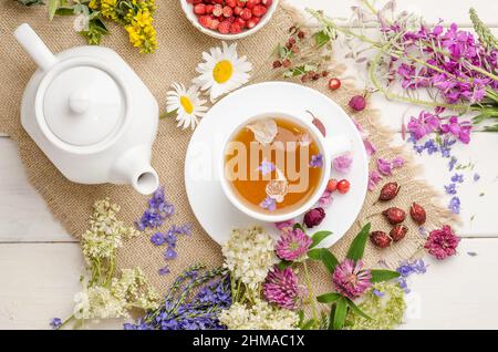 Tisane à la rosehip, camomille et limbes dans une tasse blanche sur une table blanche en bois avec fleurs, plat Banque D'Images