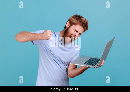 Portrait d'un homme qui frappe l'écran d'un ordinateur portable, qui regarde avec furieuse expression folle, boxe menaçant de frapper tout en ayant une conversation en ligne, appel vidéo. Studio d'intérieur isolé sur fond bleu. Banque D'Images