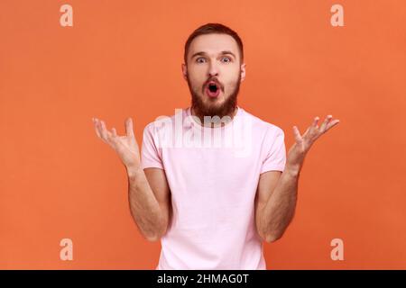 Portrait de l'homme demandant ce que vous voulez et en écartant les mains, en regardant sérieusement l'appareil photo, querelle familiale, malentendu, portant un T-shirt rose. Studio d'intérieur isolé sur fond orange. Banque D'Images