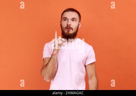 Portrait d'un beau barbu inspiré homme pointant du doigt vers le haut et regardant stupéfait de génie soudain idée, a la solution, portant le T-shirt rose. Studio d'intérieur isolé sur fond orange. Banque D'Images