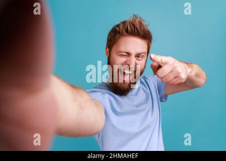 Portrait de l'homme barbu heureux heureux blogueur pointant vers vous, regardant l'appareil photo avec le sourire, POV, point de vue de la photo. Studio d'intérieur isolé sur fond bleu. Banque D'Images