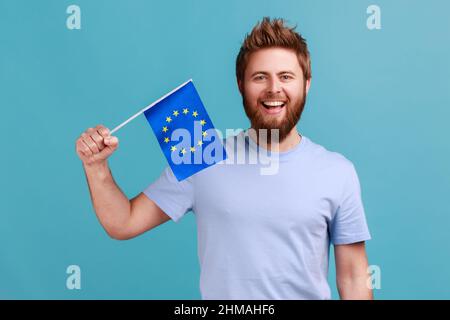 Portrait d'un homme barbu excité souriant largement et portant le drapeau de l'Union européenne, symbole de l'Europe, de l'association et de la communauté de l'UE. Studio d'intérieur isolé sur fond bleu. Banque D'Images