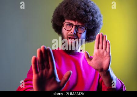 Hipster homme avec la coiffure afro montrant stop geste avec sa paume, étant contre l'intimidation et la violence, portant un sweat-shirt rouge. Studio d'intérieur isolé sur fond de néon coloré. Banque D'Images