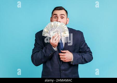 Homme d'affaires portant un costume sombre de style officiel cachant la moitié de la face derrière un fan de billets de cent dollars, choqué par le grand gagnant, pariant. Studio d'intérieur isolé sur fond bleu. Banque D'Images