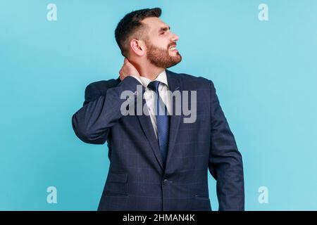 Portrait d'un homme barbu inquiet et anxieux portant un costume officiel touchant le cou, souffrant de douleurs aiguës en mouvement et en rotation de la tête, souffrant de problèmes de colonne vertébrale. Studio d'intérieur isolé sur fond bleu. Banque D'Images