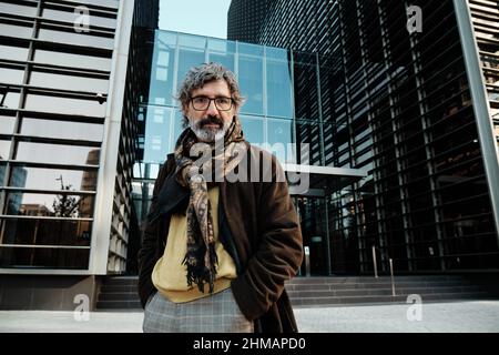 Homme d'âge moyen regardant la caméra tout en posant dans le quartier financier avec un immeuble de bureaux en arrière-plan. Banque D'Images