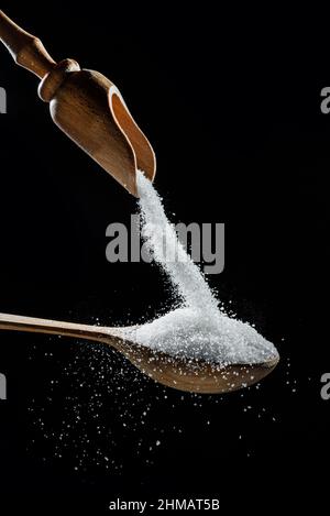 Cristaux de sucre blanc raffiné tombant dans la cuillère en bois sur fond noir. Banque D'Images