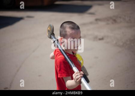 L'enfant porte un scooter. Garçon tire le scooter autour de son cou. Poids de levage de l'outil de préchoix. Un jeune athlète porte de l'équipement sportif. Enfant en T-shirt rouge. Mon Banque D'Images