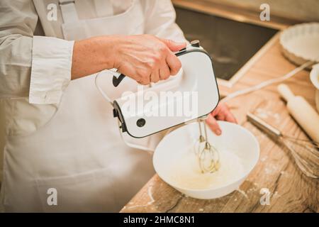 Femme adulte en blanc de vêtements de chef bat des oeufs avec le mélangeur pour faire la tarte dans la cuisine maison Banque D'Images