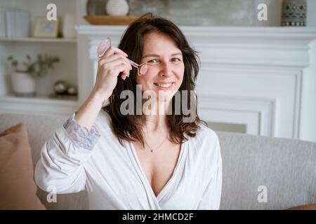 Brunette femme souriante de 35 ans, visage et mains propres et frais avec des cheveux longs, fait un massage du visage avec un rouleau de gouache dans un peignoir tout en étant assis à la maison sur le canapé.Concept de soin du visage Banque D'Images