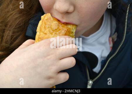 gros plan de la bouche de la jeune fille en prenant une bouchée d'un en-cas de pâtisserie. La main de l'enfant tient le rouleau de saucisse le mettant sur ses lèvres, à l'extérieur Banque D'Images