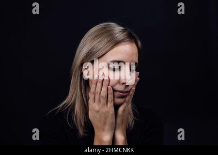 Femme déprimé visage avec les mains touchant les joues portant une robe noire sur fond noir. Victime d'abus physique et psychologique. Éclairage au gaz Banque D'Images