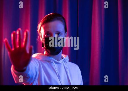 Jeune belle femme dans un masque médical noir et un chandail à capuchon blanc sur fond bleu-violet montre des gestes de main pendant une pandémie de coronavirus Banque D'Images