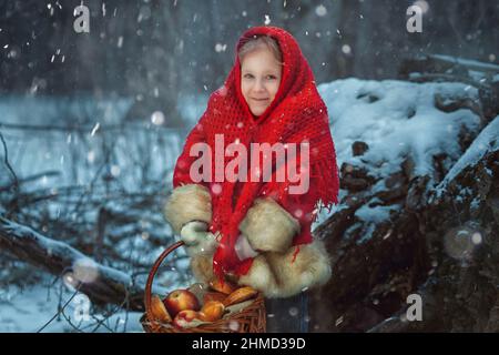 Portrait d'une petite fille dans la forêt d'hiver. Elle a un panier de pommes et de tartes entre ses mains. Banque D'Images