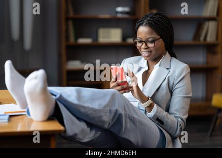 Une femme africaine heureuse assise avec les jambes au bureau défilant sur les réseaux sociaux sur un smartphone Banque D'Images