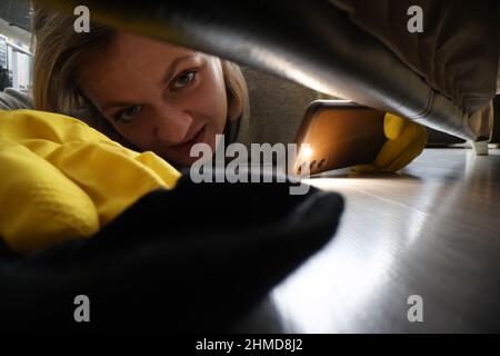 Femme à la recherche d'une chaussette sous le canapé à l'aide d'une lampe de poche sur le téléphone portable Banque D'Images