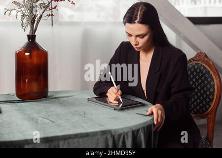 Jeune femme freelance brune designer dessine des croquis sur une tablette graphique à l'aide d'un stylet. Habillé d'un élégant costume noir. Intérieur classique Banque D'Images