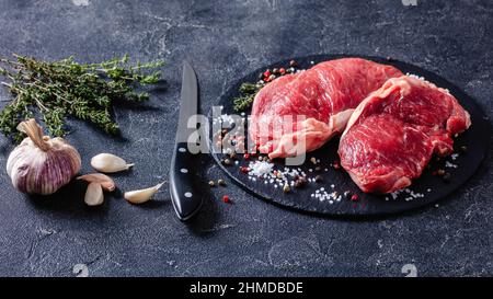 Steaks de New York crus sur une assiette en pierre noire avec sel et poivre sur une table de cuisine en béton Banque D'Images