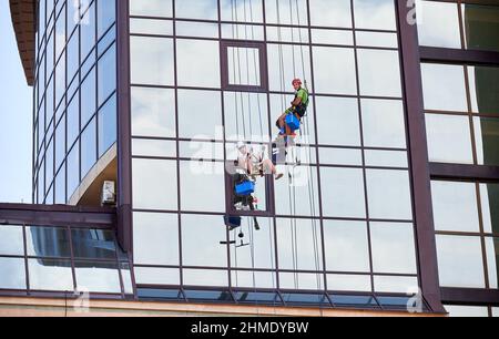 Ouvriers d'alpinisme industriel lavant des fenêtres en verre d'une tour de bâtiment, accrochés à des cordes d'escalade de sécurité. Deux nettoyeurs de fenêtre hommes travaillant ensemble à l'extérieur du gratte-ciel. Banque D'Images