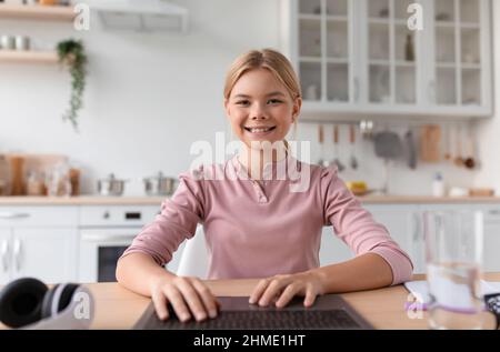 Portrait d'une jeune fille blonde blanche souriante tapant au clavier par ordinateur portable dans la cuisine intérieure Banque D'Images